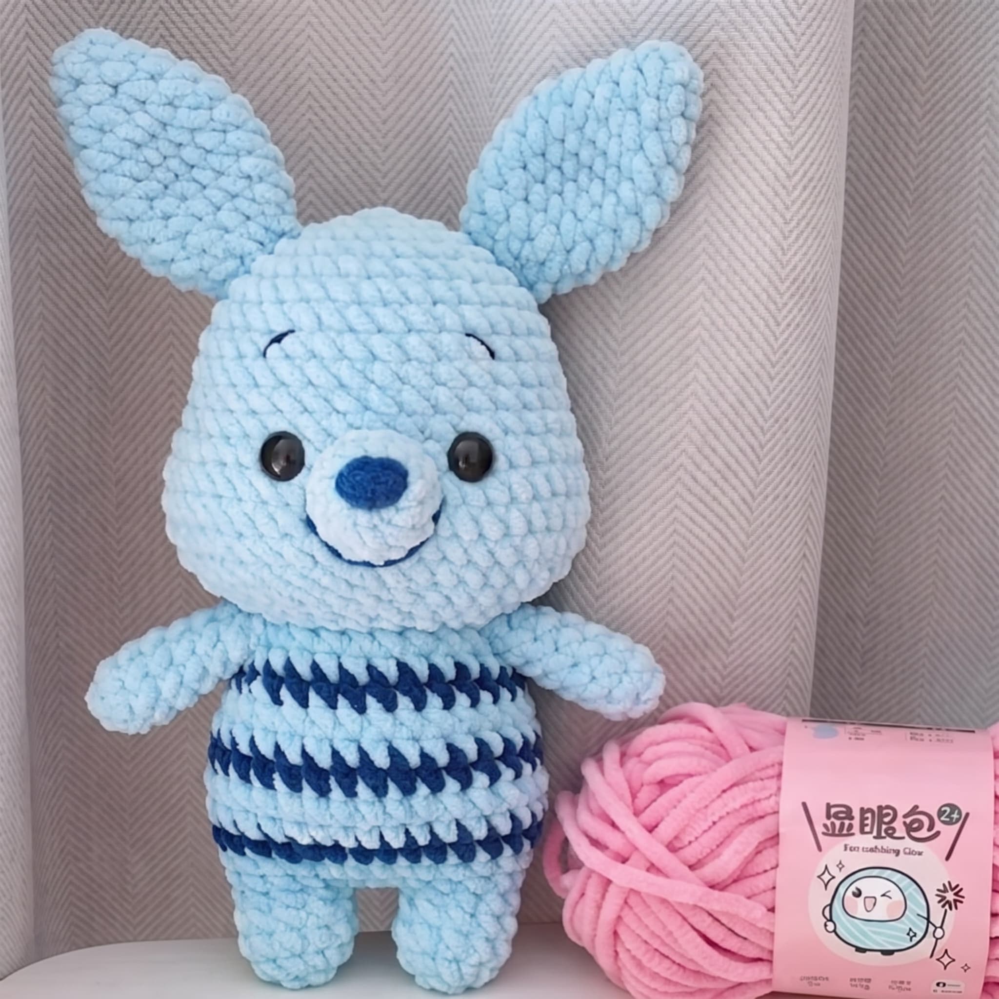 Blue plush crochet bunny amigurumi with a navy-striped belly beside a ball of pink chenille yarn