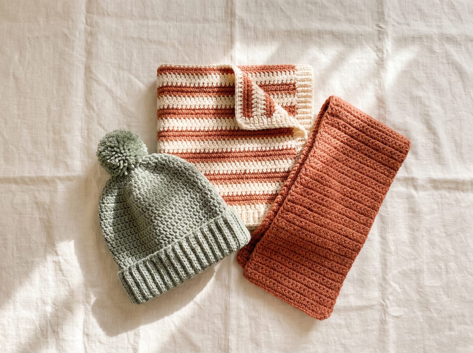 Three finished half double crochet projects, a beanie, a baby blanket, and a scarf, arranged on linen.