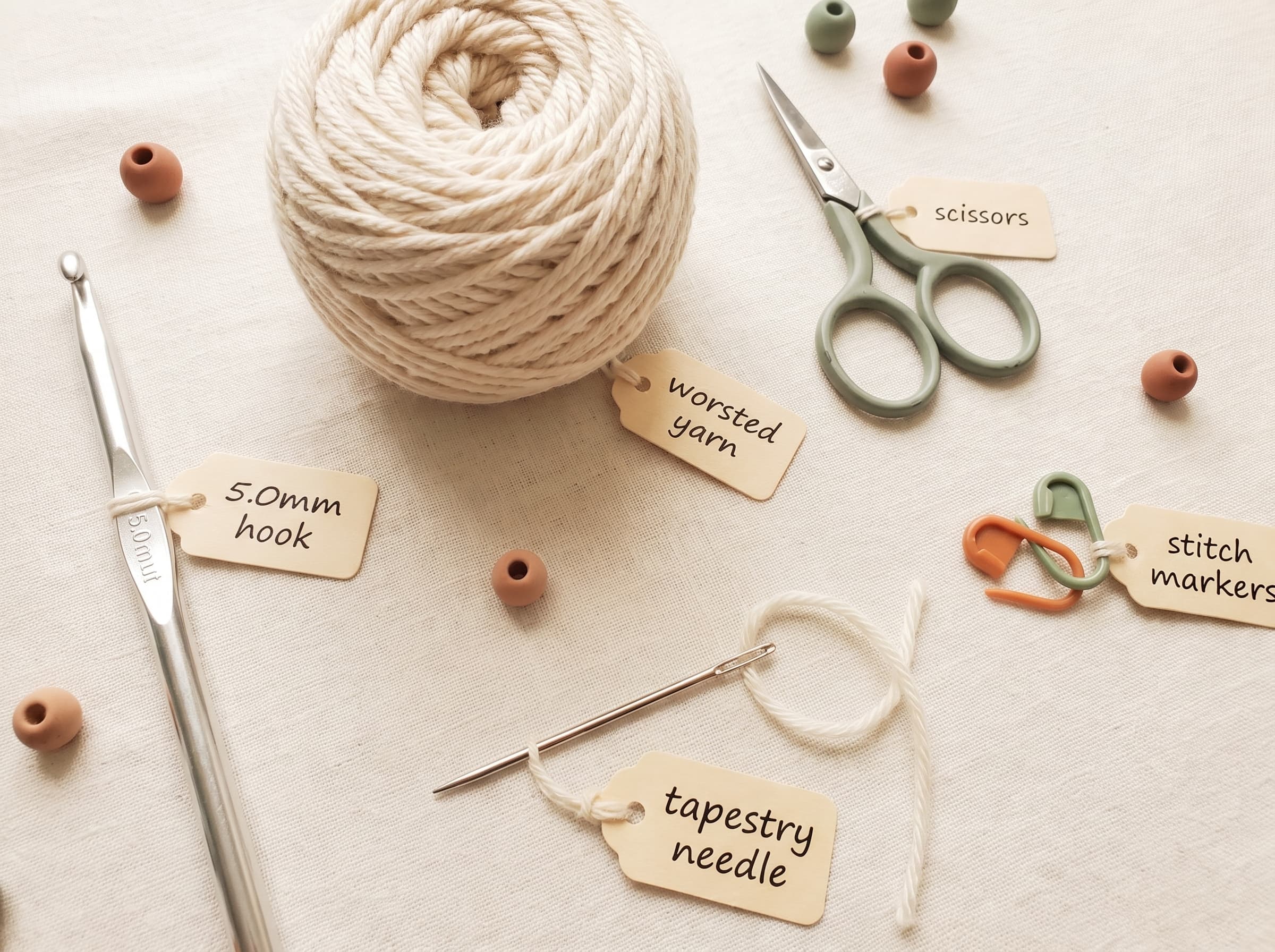 Flat-lay of beginner crochet tools labeled: 5.0mm hook, worsted yarn, scissors, tapestry needle, and stitch markers.