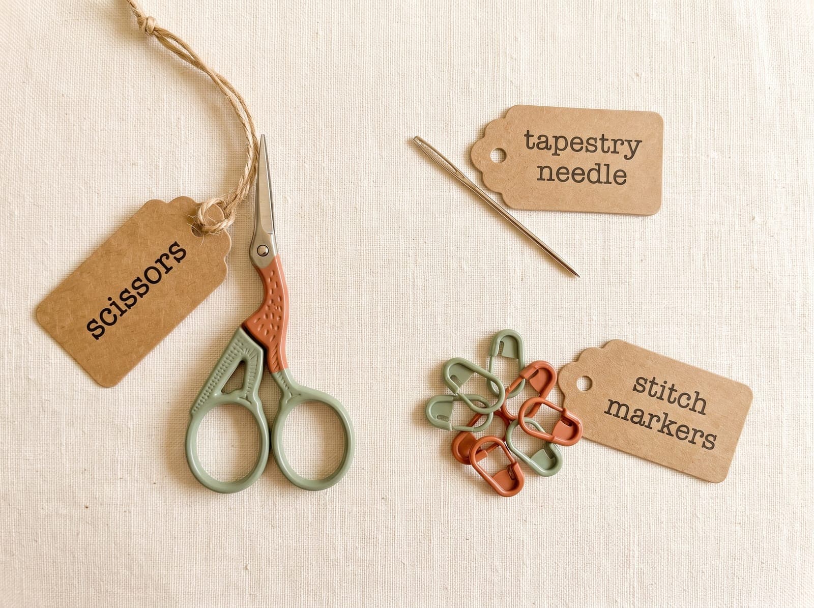 Flat-lay of crochet notions labeled scissors, tapestry needle, and stitch markers on cream linen.