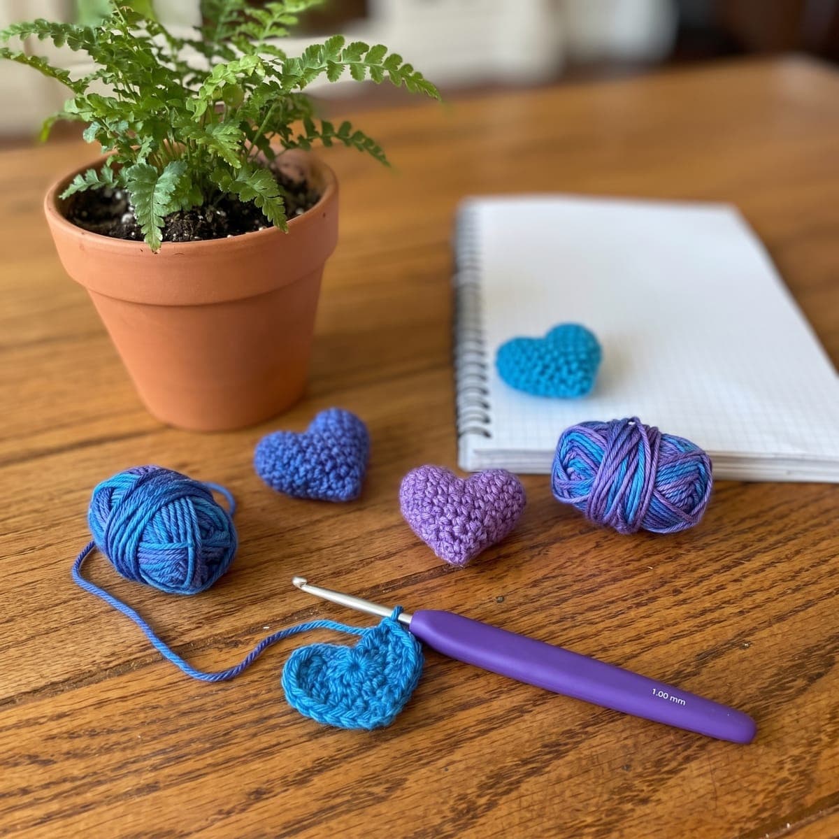 Crochet hearts in blue and purple yarn beside yarn balls, a crochet hook and a notebook on a wooden table.